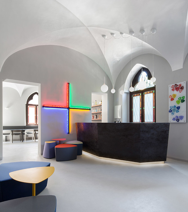Modern reception area in vaulted space with Artemide globe pendant lights and colorful neon cross wall installation above dark reception desk.