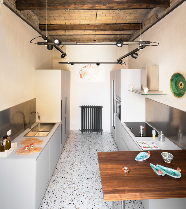 Modern kitchen with Artemide track lighting system featuring adjustable black spotlights suspended from exposed wooden beams over terrazzo floor.