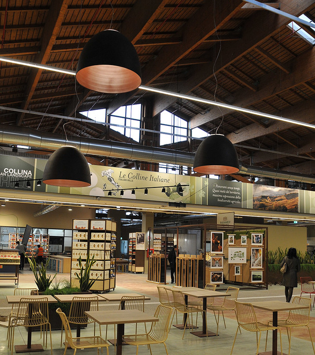 Large black pendant lamps with copper interior illuminate a spacious Italian food market hall with wooden ceiling and dining area.