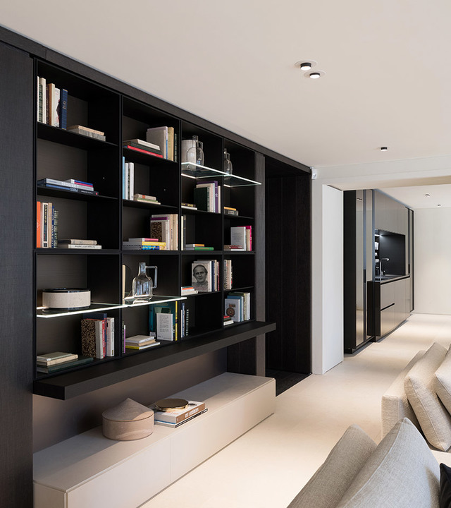 Modern living room with dark built-in bookshelf lit by integrated LED strip lights, ceiling spotlights, and light-toned sofa nearby.