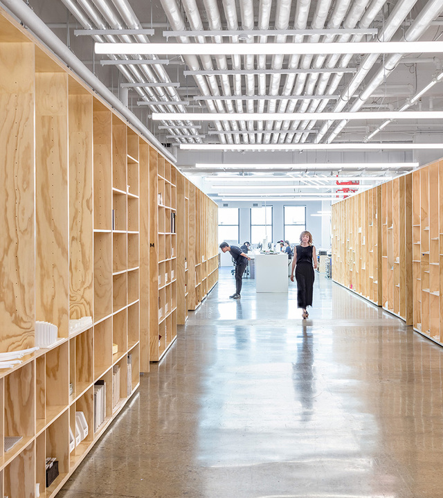 Modern office corridor with plywood shelving walls, polished concrete floor, and linear LED ceiling lights illuminating the workspace.