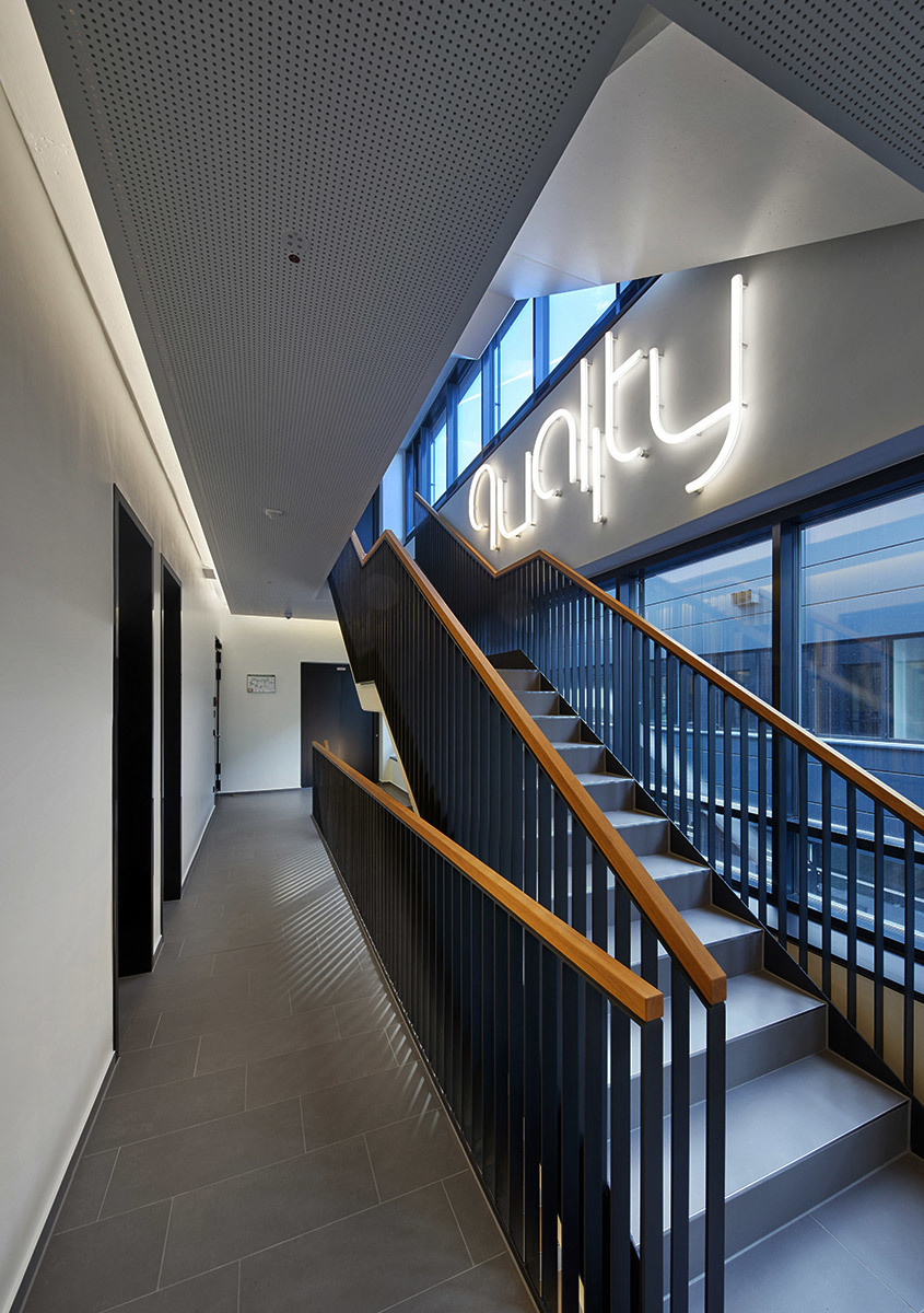 Modern office stairwell with dark metal railings, wood handrails, and a glowing neon 'quality' sign mounted on the wall.