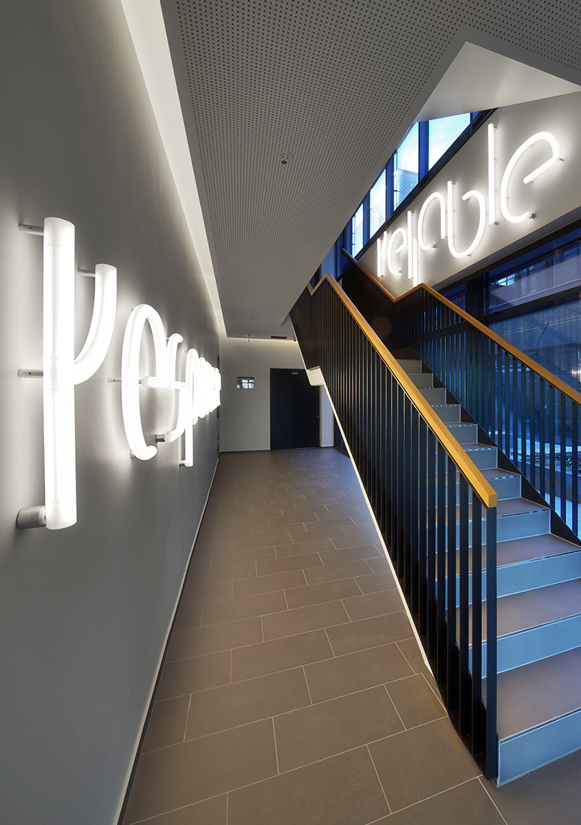 Modern corridor with illuminated 3D letter signs on walls spelling 'people' and 'reliable', with recessed ceiling lights and a staircase.