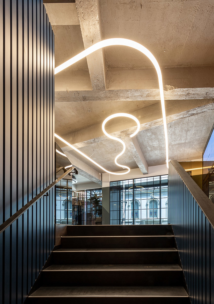 Artemide flexible LED light system forming abstract curves on exposed concrete ceiling above a modern staircase with dark metal railings