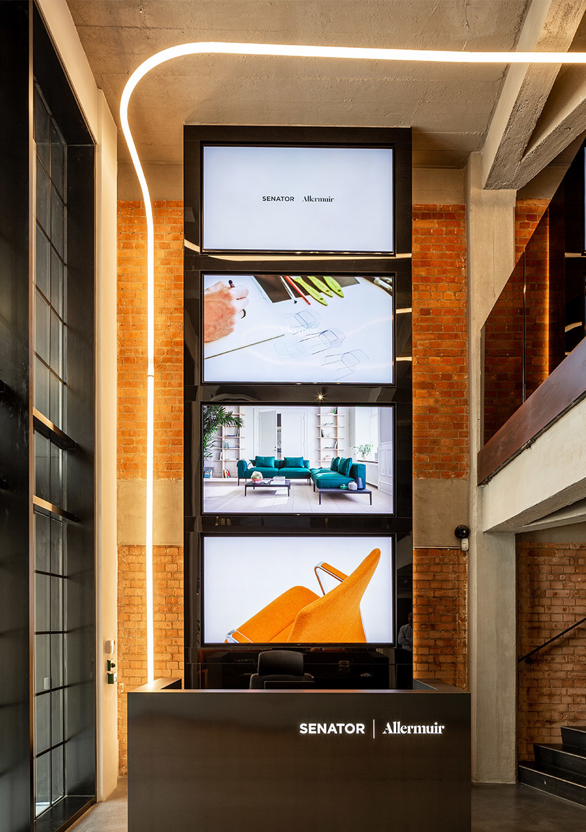 Senator and Allermuir branded display tower with stacked screens showing furniture collections, illuminated by a curved LED strip light.
