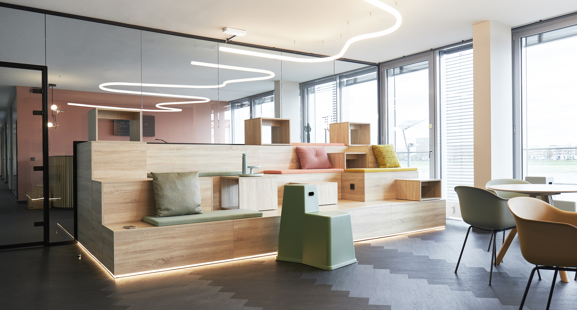 Modern office lounge with Artemide curved LED ring pendant lights illuminating wooden modular seating and colorful cushions.