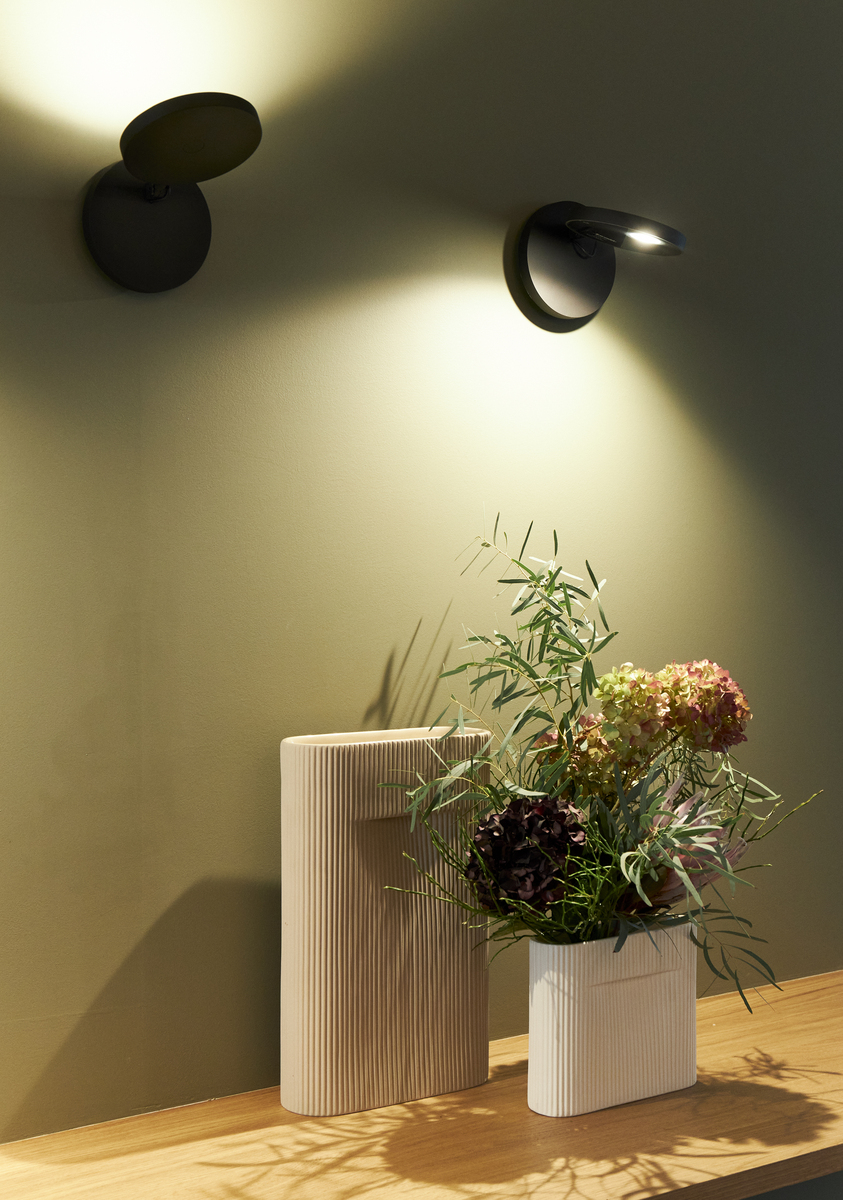 Two black wall-mounted adjustable spotlights casting warm light on a shelf with ribbed vases and a floral arrangement.