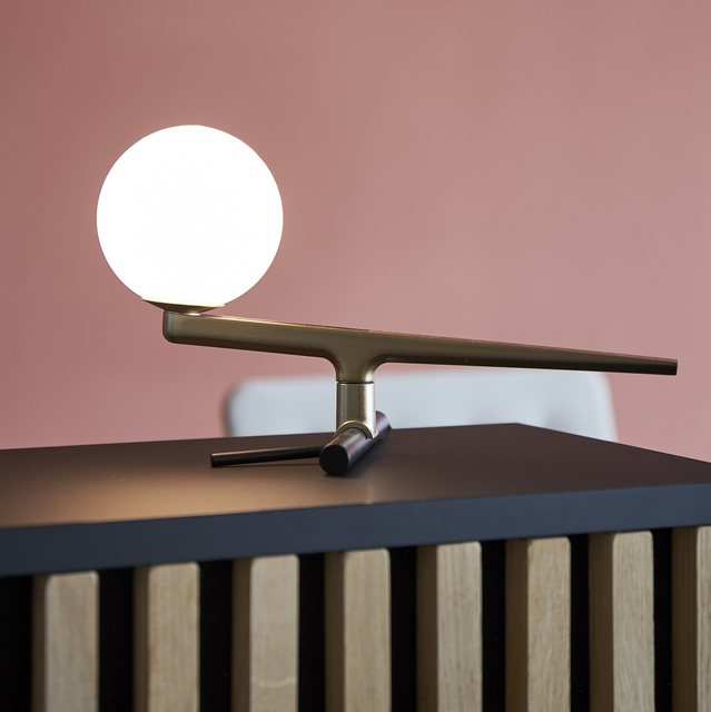 Artemide table lamp with brass horizontal arm, white opal glass sphere, and black vertical base, placed on a dark sideboard.