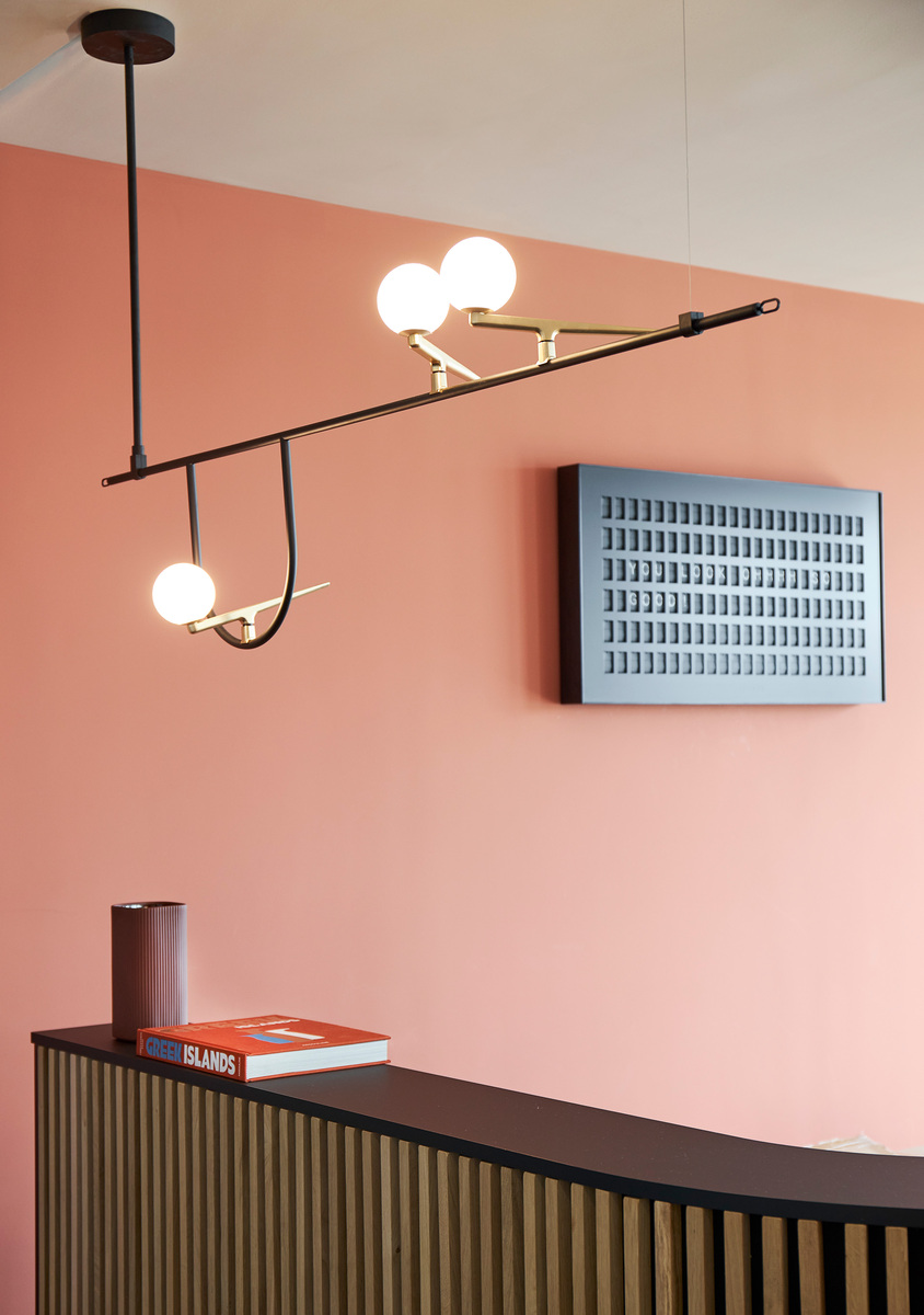 Artemide pendant lamp with black arms and brass joints holding three opal glass spheres, installed above a reception desk on a pink wall.