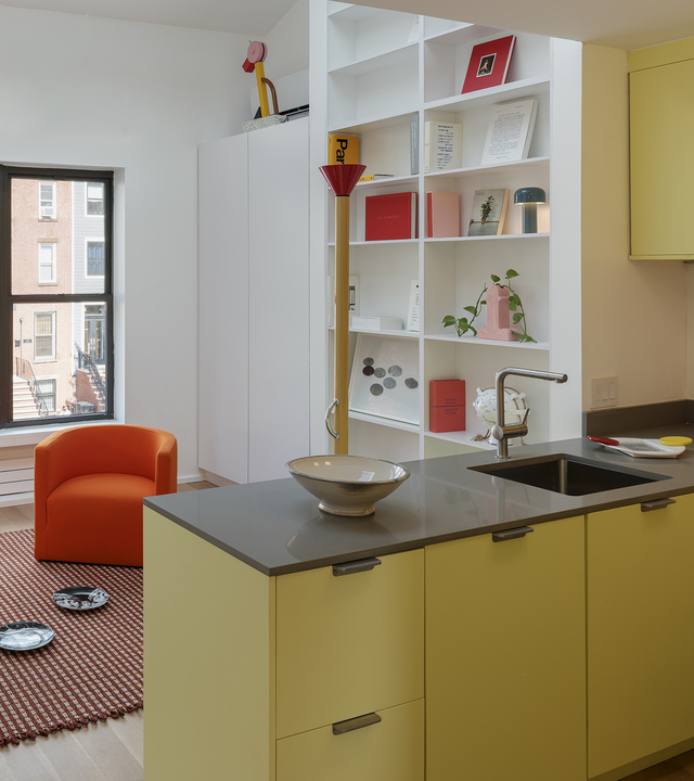 Modern kitchen with yellow cabinets and gray countertop, open shelving unit displaying colorful books and a pink table lamp