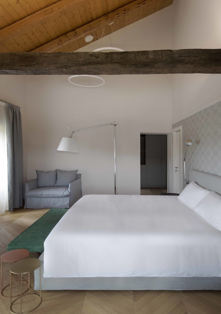 Hotel bedroom with Artemide Tolomeo floor lamp beside a gray armchair, white bed, herringbone wood floor, and vaulted wooden ceiling