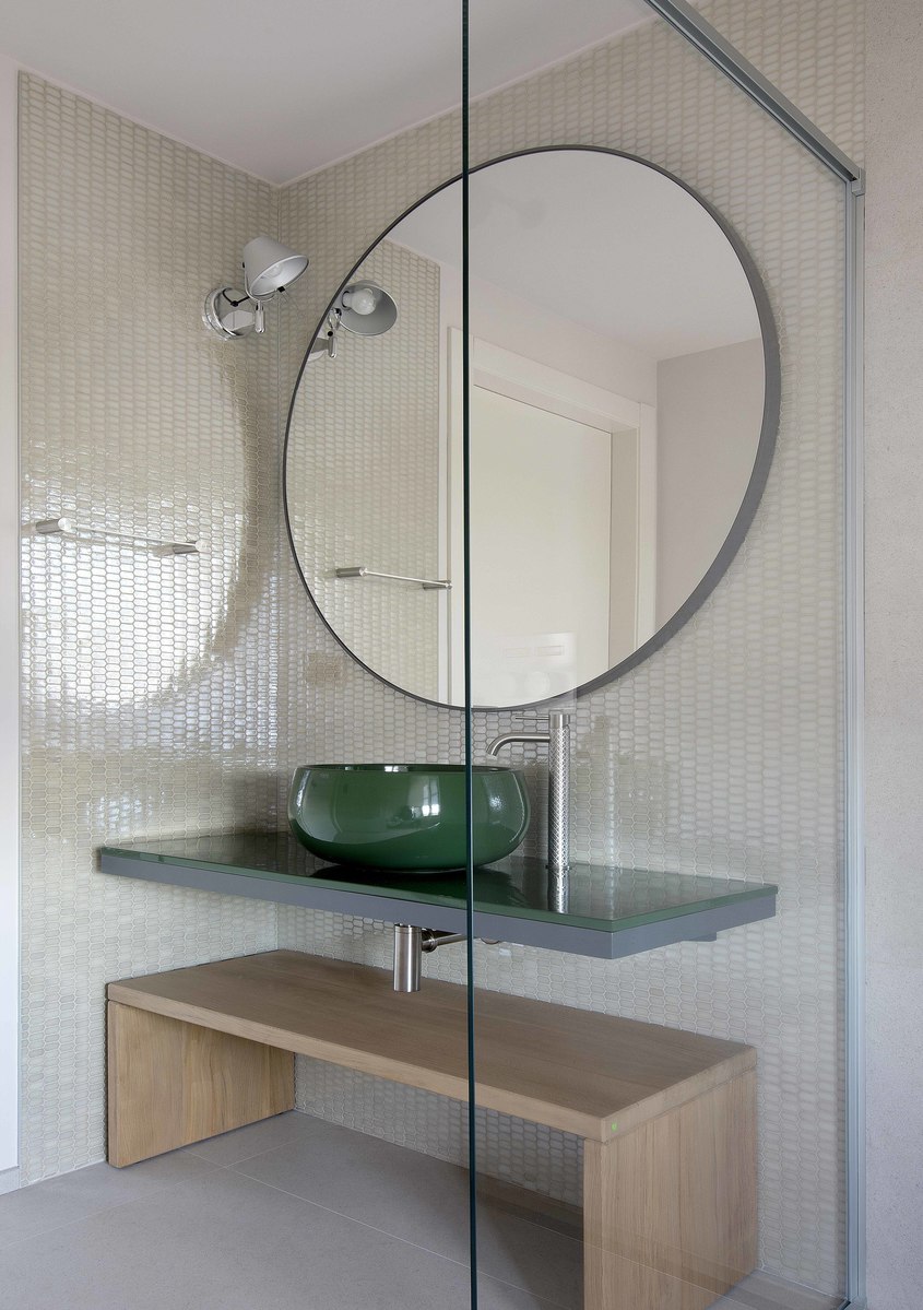 Modern bathroom with Artemide wall lamp, round mirror, green vessel sink on floating shelf, and wooden bench
