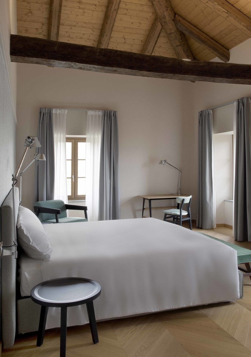Elegant hotel bedroom with wooden beam ceiling, white bedding, small black stool, and two Artemide Tolomeo desk lamps on bedside and desk.