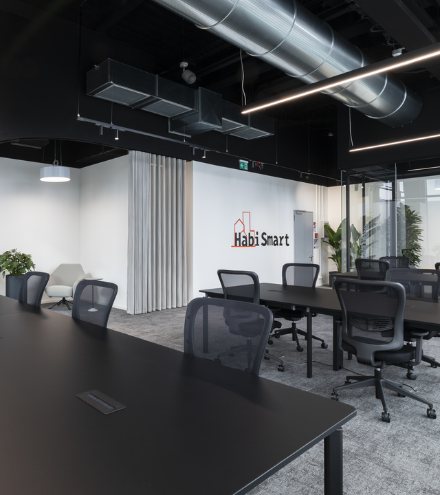 Modern office with black desks, mesh chairs, exposed ductwork ceiling, linear LED pendant lights, and HabiSmart branding on white wall.