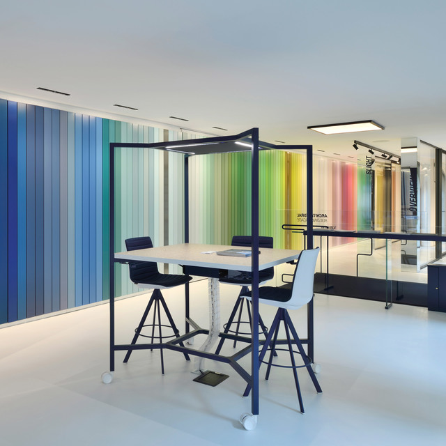 Modern workspace with high table, bar stools under a black frame canopy, and recessed ceiling lights beside a vibrant multicolored striped wall.