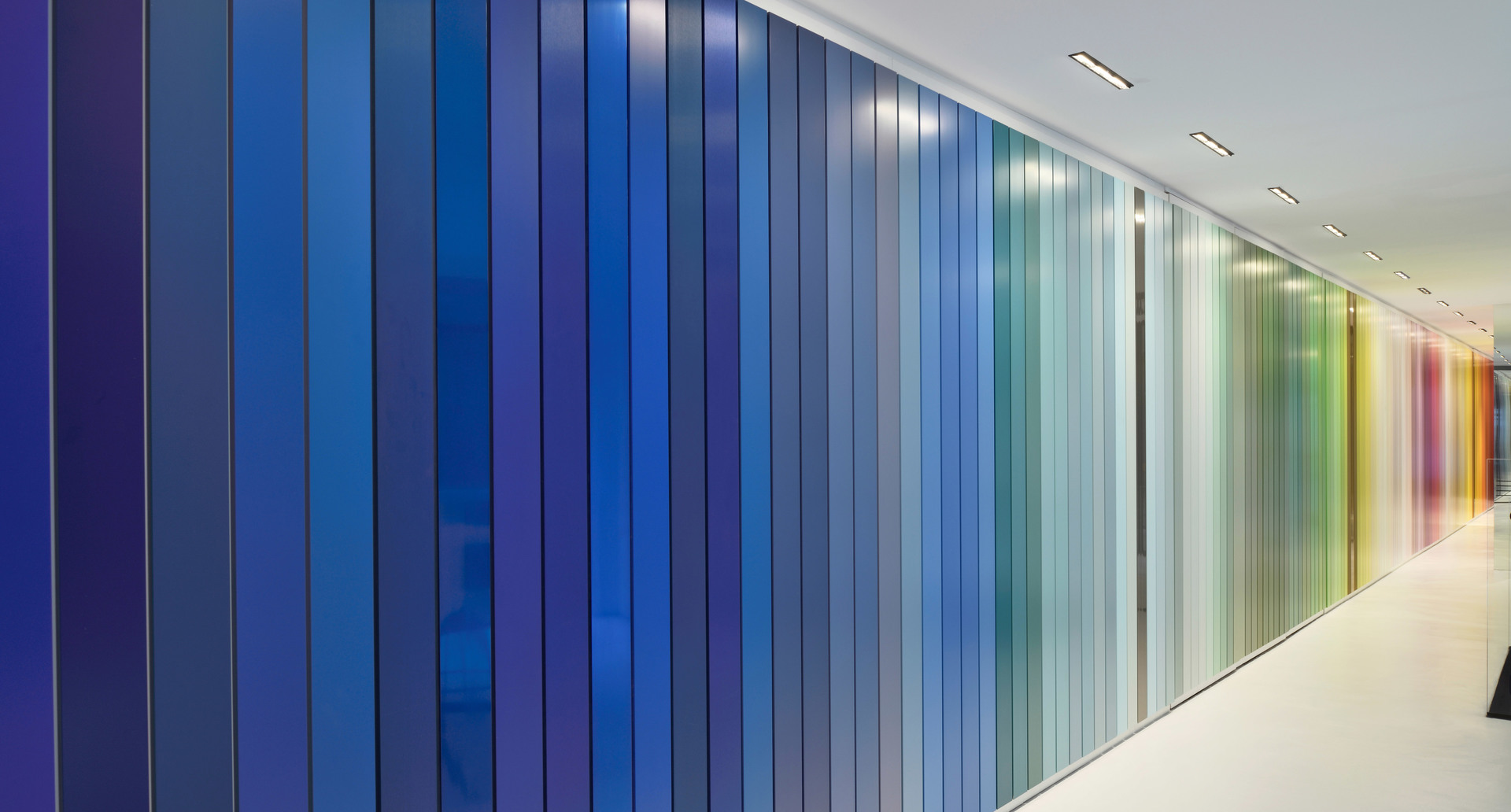 Corridor with a vibrant floor-to-ceiling color gradient wall, transitioning from deep blue to green and warm tones, lit by recessed ceiling lights.