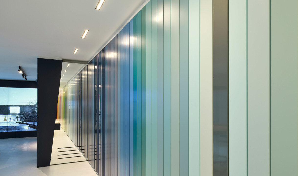 Modern corridor with a large illuminated wall of vertical multicolored glass panels, transitioning from blue to green tones, lit by recessed ceiling lights.