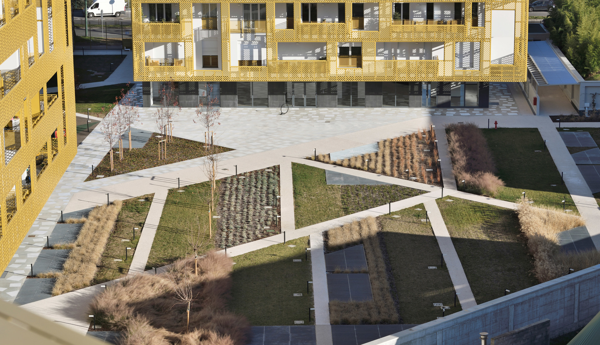 Aerial view of a modern courtyard with geometric green areas, pathways, young trees, and low bollard lights between yellow-facade buildings.