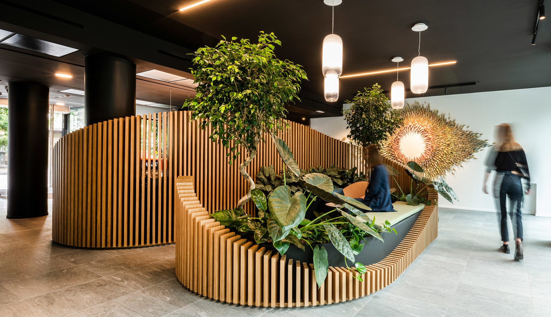 Modern office lobby with curved wooden slatted seating, lush greenery, and Artemide cylindrical pendant lights on dark ceiling