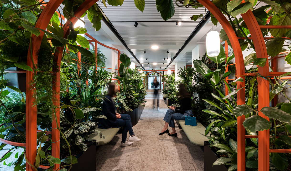 Office corridor with orange arched frames, lush tropical plants, seating areas, and white pendant lights overhead.