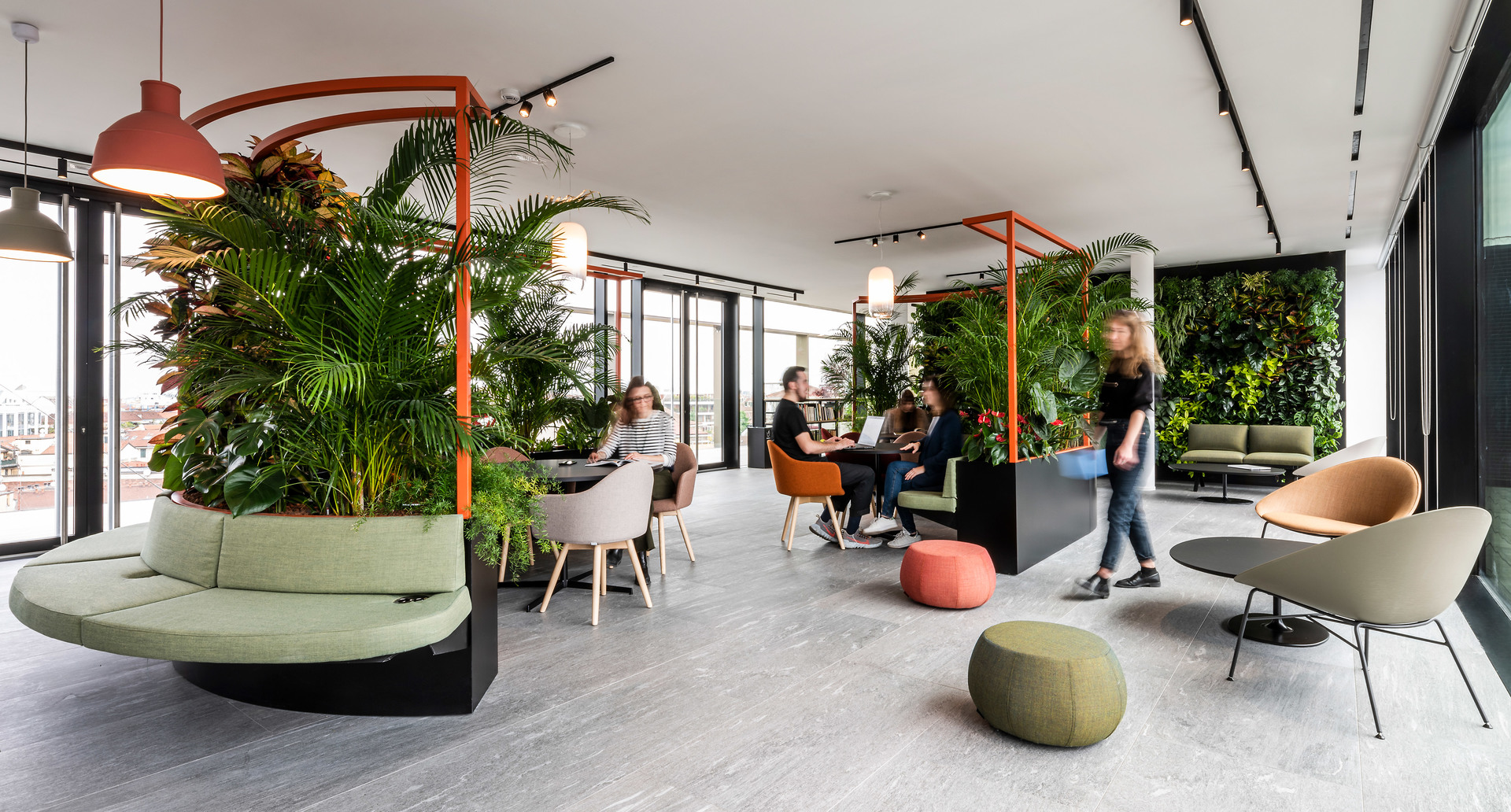 Modern biophilic office lounge with lush greenery, orange structural frames, track lighting, and collaborative seating areas where people work and socialize