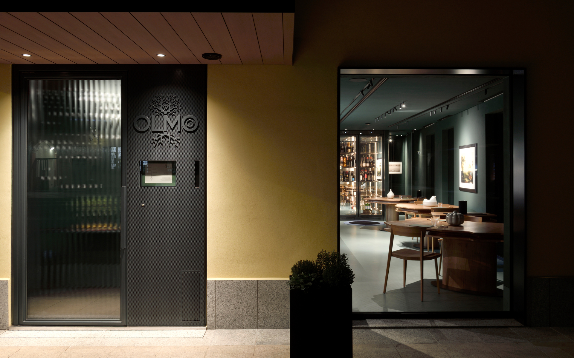 Olmo restaurant entrance at night, warm-lit interior visible through glass door with track lighting illuminating dining area