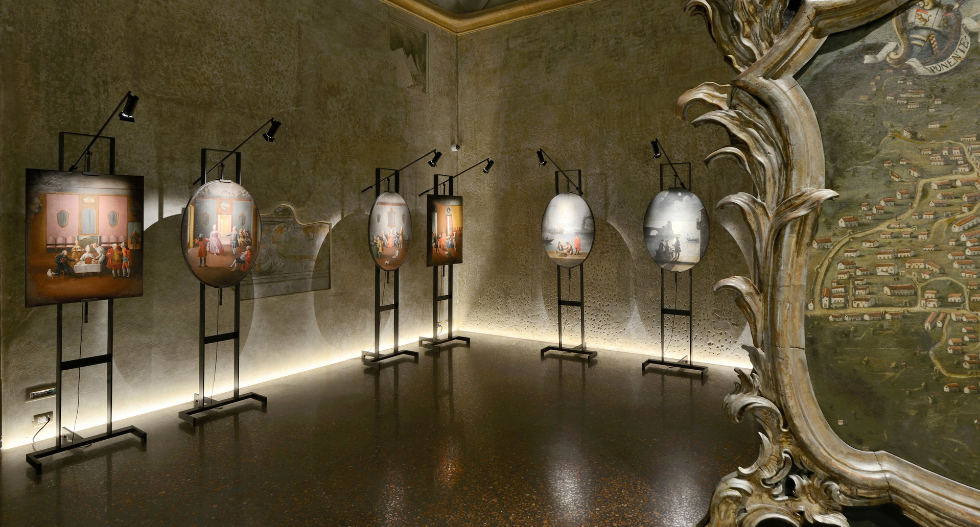 Gallery room with artworks on easels lit by adjustable black spotlights, warm LED strip lighting along the base of textured walls.