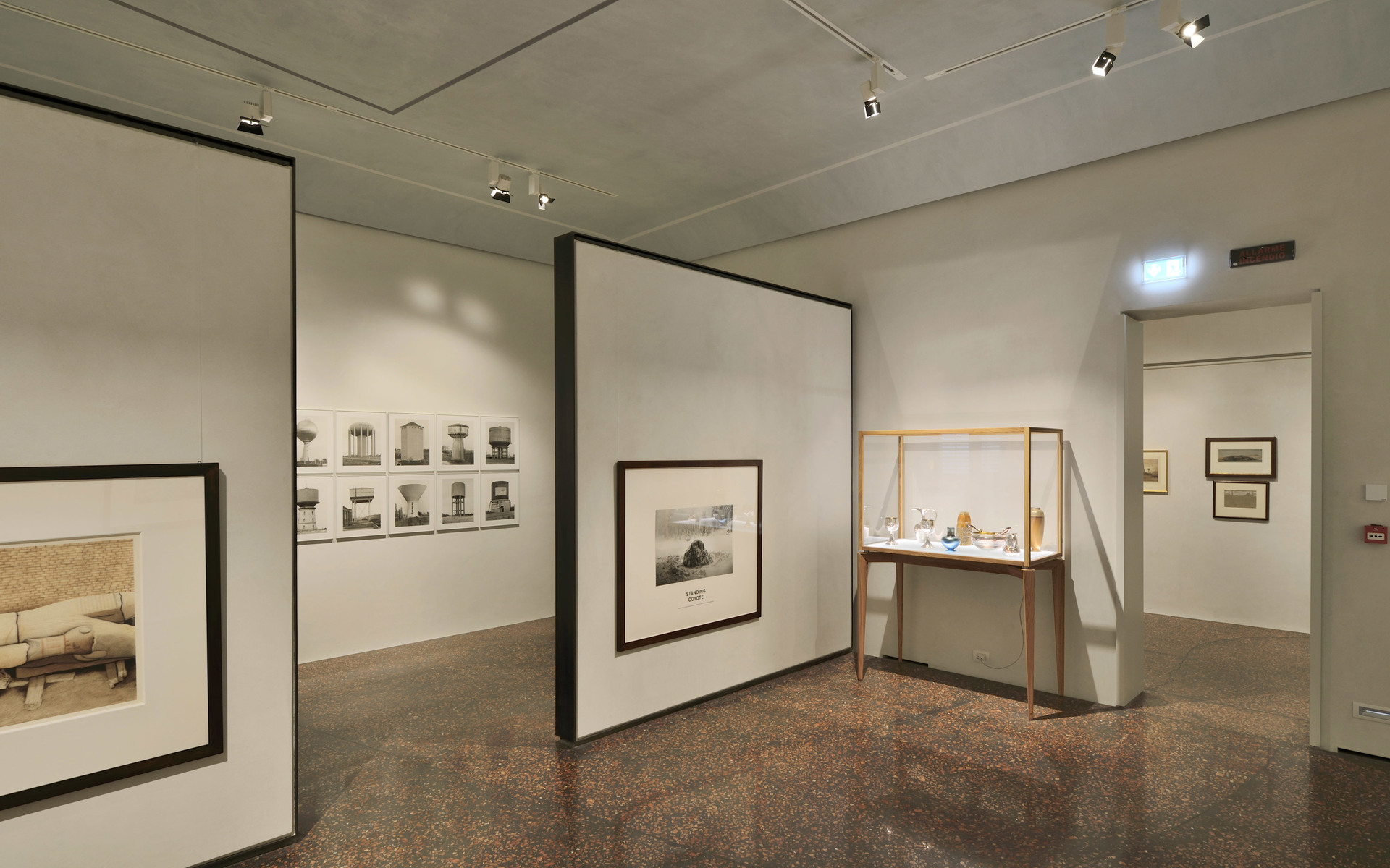 Museum gallery with Artemide ceiling spotlights illuminating framed artworks on white walls and a glass display case with objects.