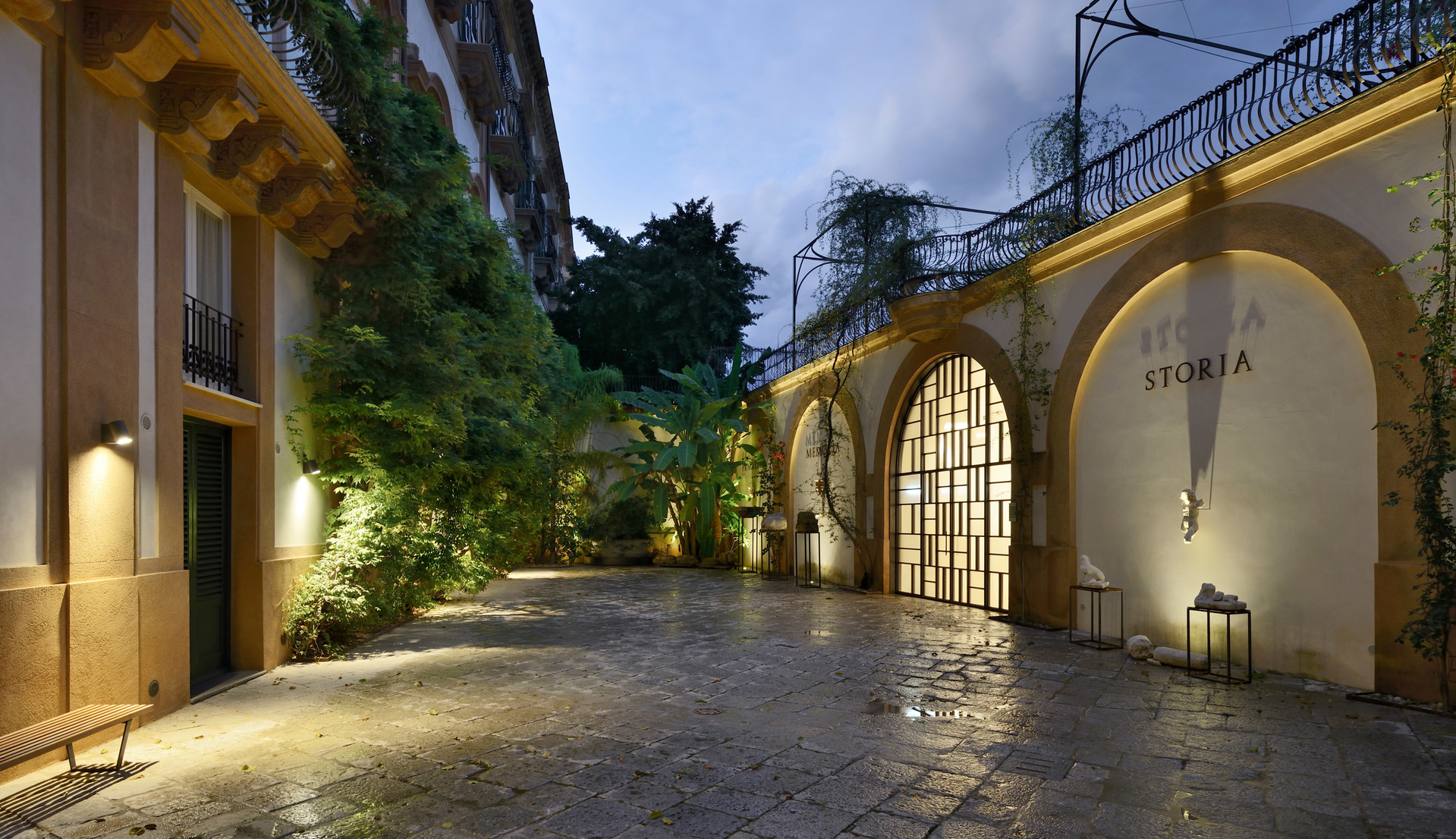 Elegant courtyard at dusk with arched facade, lush greenery, cobblestone floor, and warm Artemide wall lights illuminating the space.