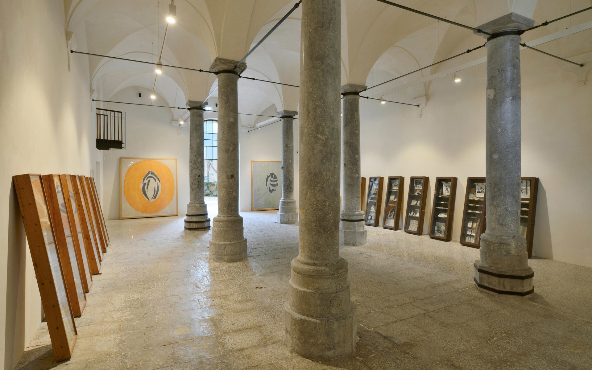 Gallery space with vaulted ceilings, stone columns, leaning wooden frames, and artworks lit by ceiling-mounted spotlights