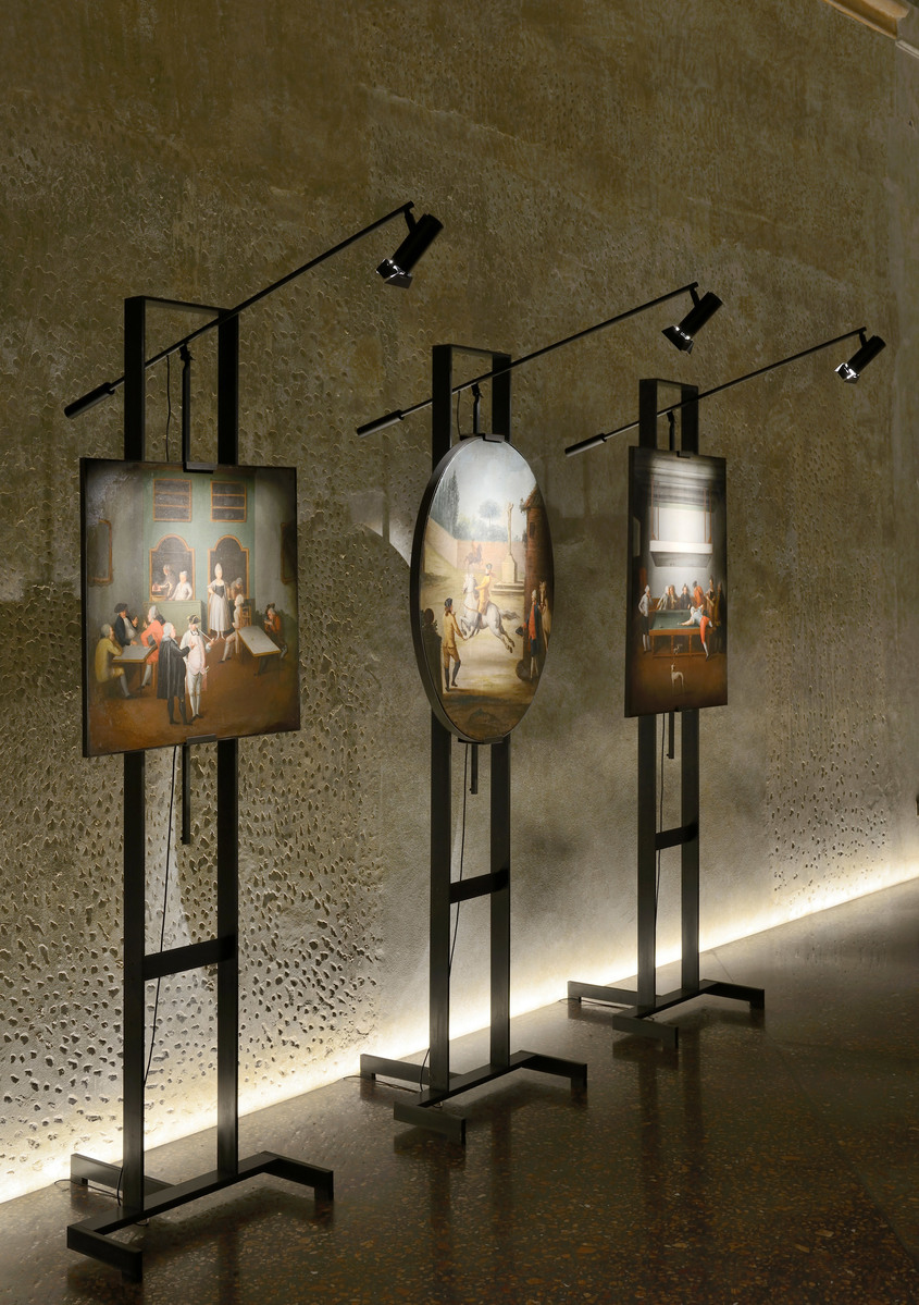 Three black metal stands displaying illuminated artworks in a gallery, each topped with an adjustable Artemide spotlight directing light onto the pieces.