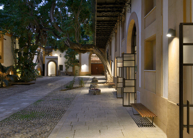 Historic courtyard at dusk with wall-mounted Artemide lights illuminating stone pathways, arched facades, and metal shelving units.