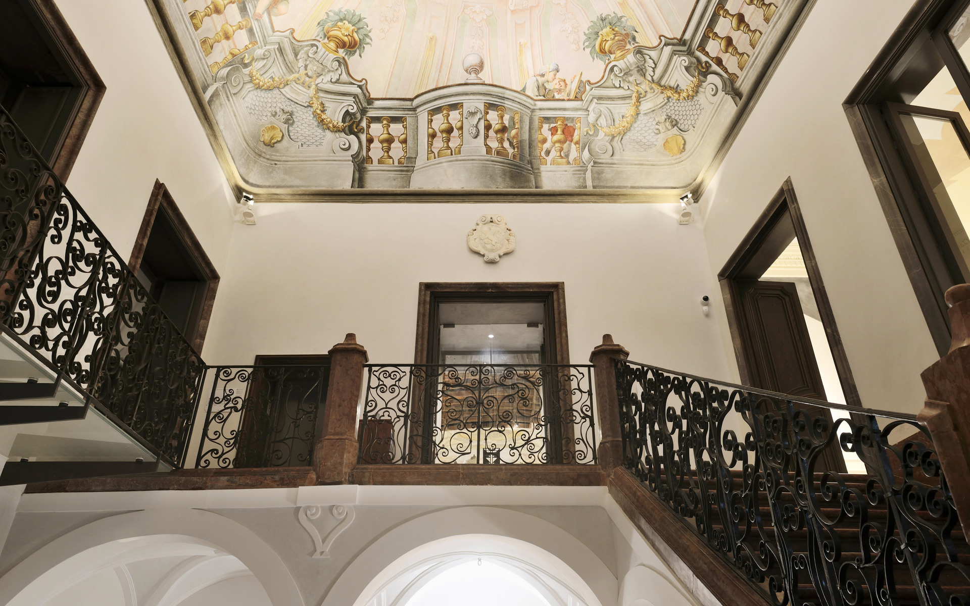 Historic palazzo interior with ornate frescoed ceiling, wrought iron balustrade staircase, and warm ambient lighting on white walls