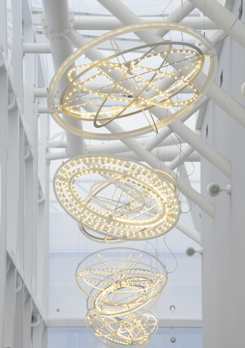 Three large Artemide pendant lights with overlapping illuminated rings in warm white LED, suspended in a bright atrium with white structural columns