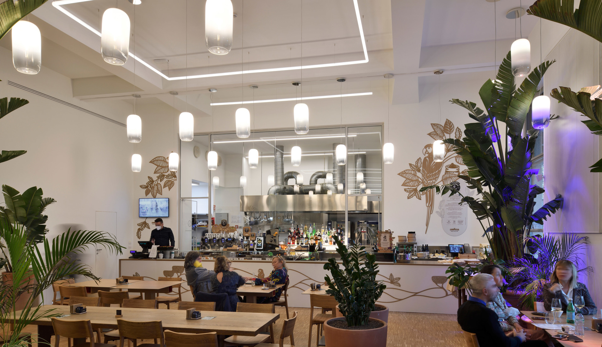 Modern café interior with multiple white cylindrical pendant lights illuminating dining area, open kitchen, and tropical plants.