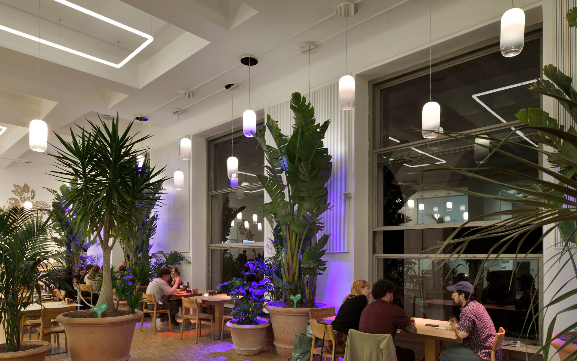 Restaurant interior with Artemide white cylindrical pendant lights, lush tropical plants, and purple accent lighting creating a warm ambiance.