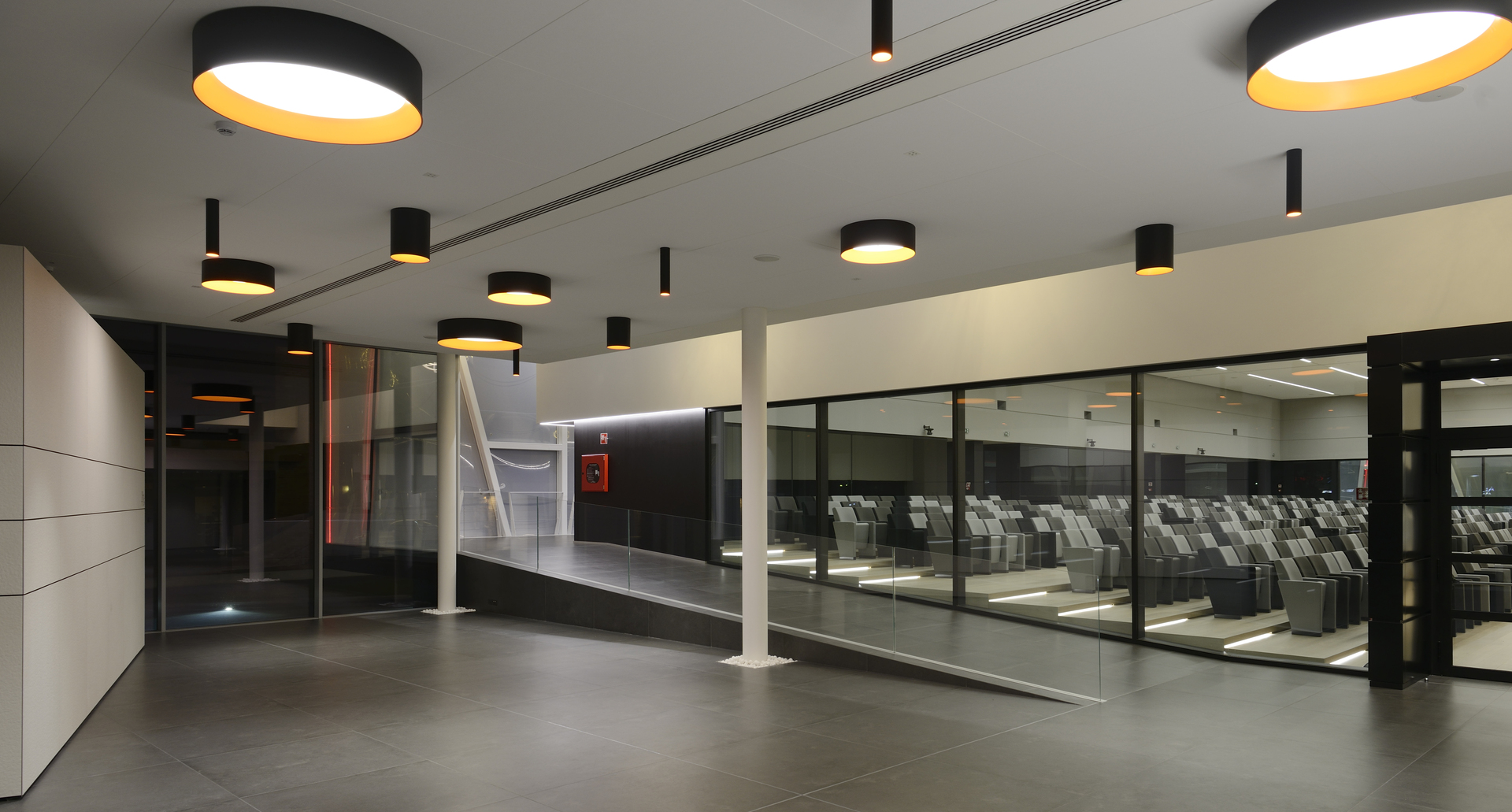 Modern lobby with large and small black cylindrical pendant lights featuring golden inner shades, illuminating a sleek open space with glass partitions.