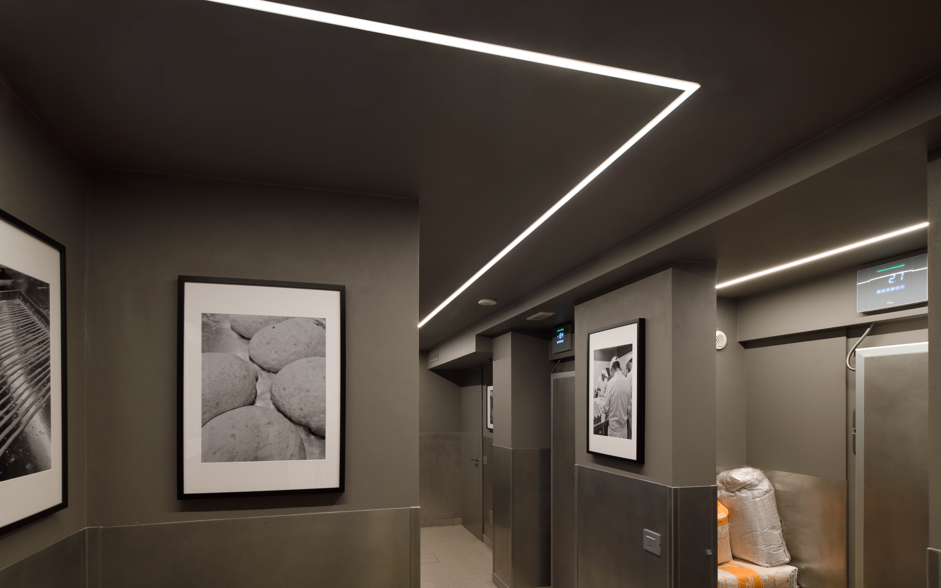 Corridor with recessed linear LED ceiling lights forming geometric angles, illuminating gray walls with black-and-white framed artwork.