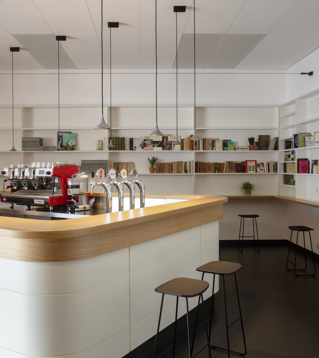 Modern café bar with curved white counter, wood top, beer taps, and Artemide pendant lights hanging from ceiling above shelved books