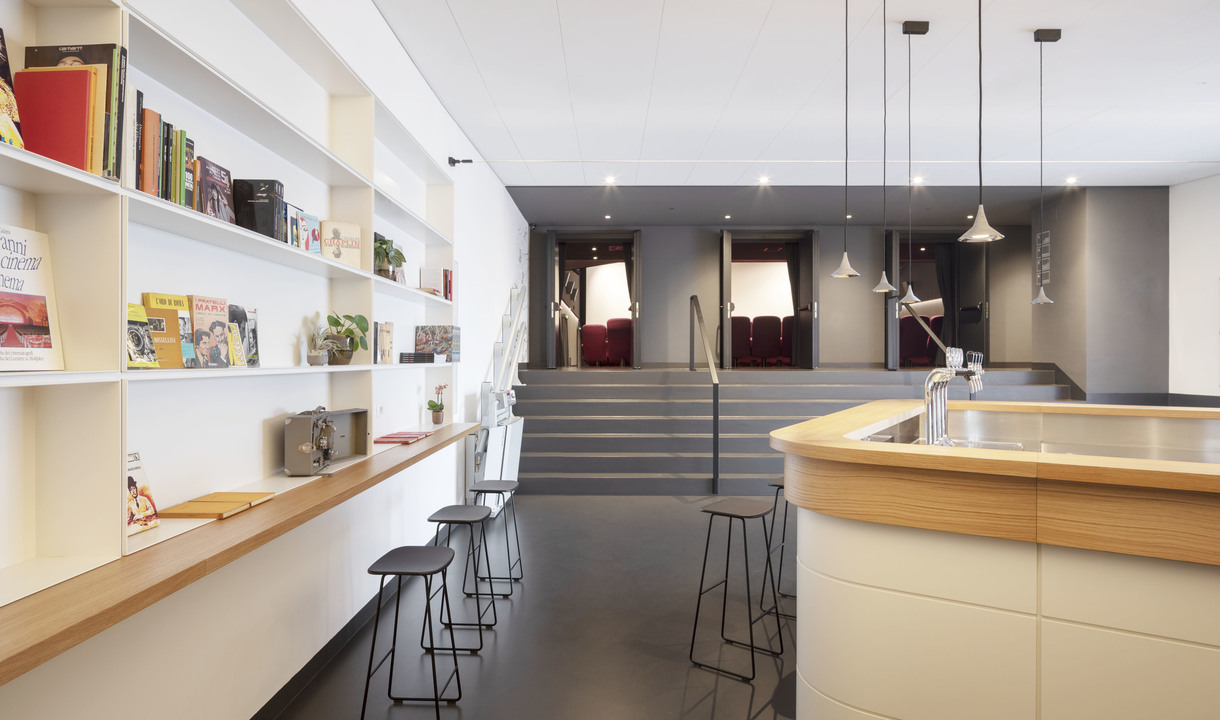 Modern lounge with Artemide pendant lights over a curved wooden counter, white bookshelves, bar stools, and steps leading to a seating area.