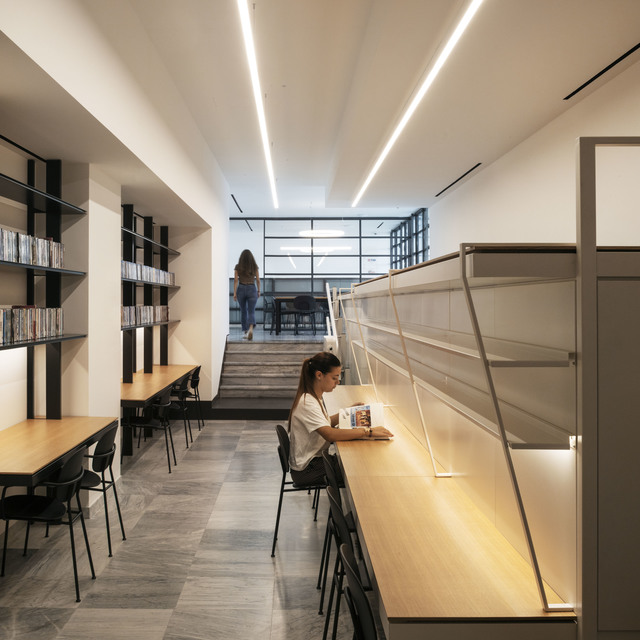 Modern library reading area with linear ceiling lights and warm task lighting illuminating a long wooden desk where a student reads.
