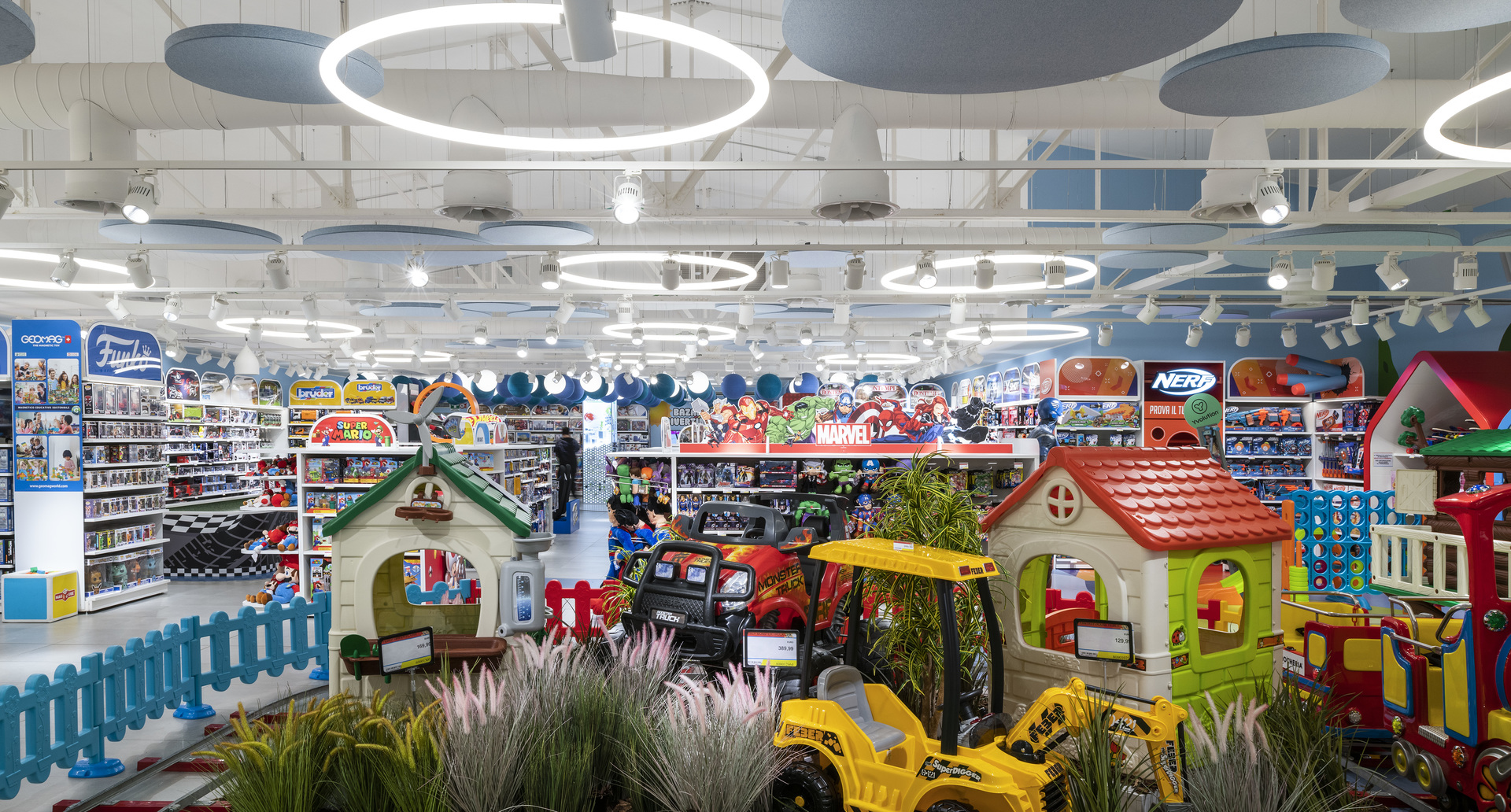 Toy store interior with circular Artemide pendant ring lights illuminating colorful displays of toys, playhouses, and ride-on vehicles.