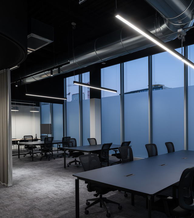 Modern open-plan office with linear pendant LED lights arranged diagonally over dark workstations, floor-to-ceiling windows at dusk.
