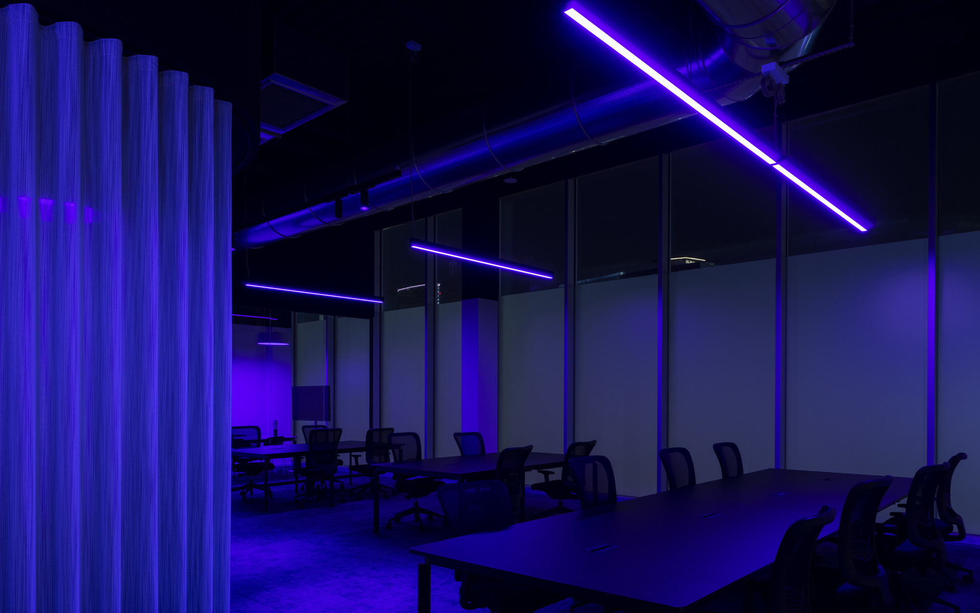 Dark office workspace with blue and violet linear LED ceiling lights creating an X-pattern, illuminating rows of desks and chairs