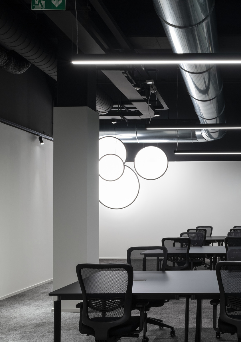 Office workspace with Artemide circular pendant lights on white wall, linear LED ceiling strips, dark tables and mesh chairs.