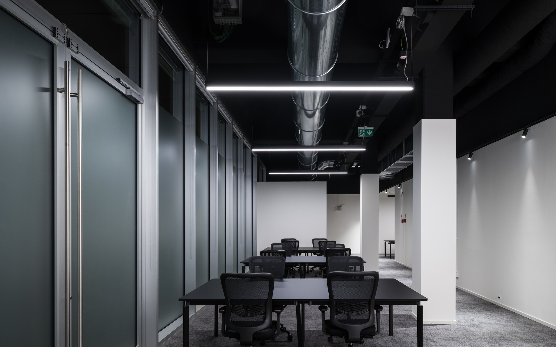Modern conference room with black ceiling, linear LED pendant lights illuminating dark tables and chairs, exposed duct, glass partition wall.