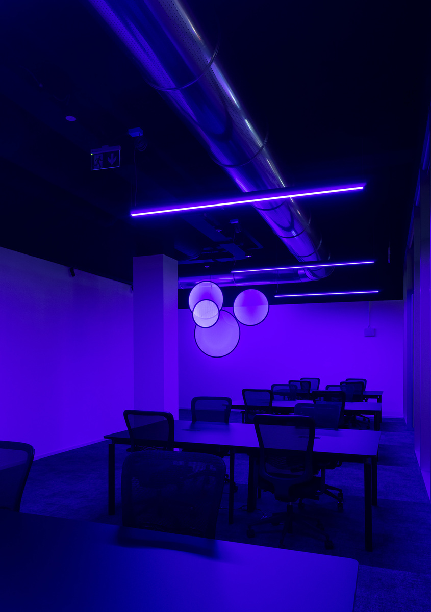 Office space bathed in purple light with linear ceiling fixtures and three spherical pendant lamps glowing on a dark wall.