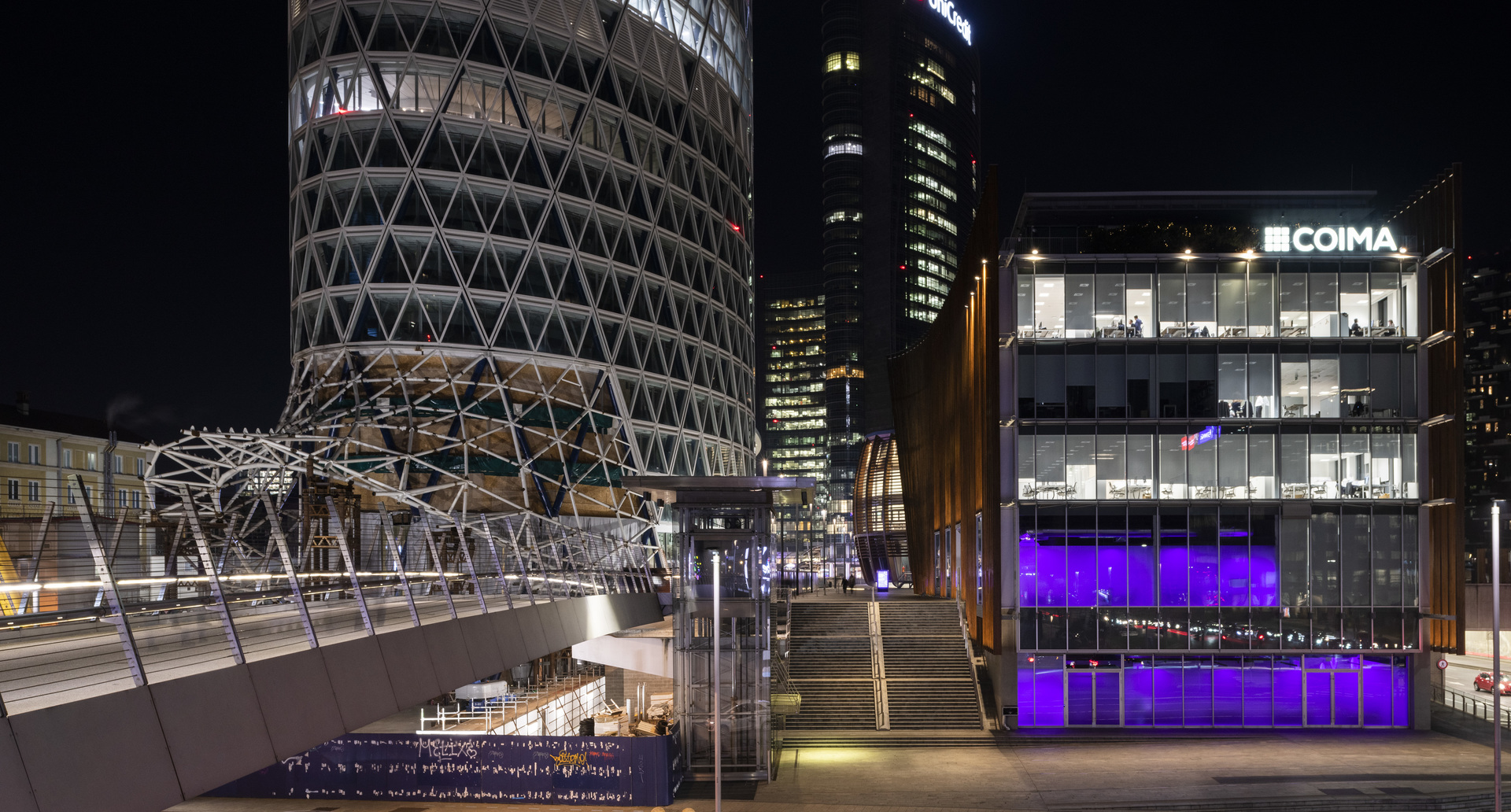 Milan cityscape at night: illuminated COIMA building with purple lighting, cylindrical tower with geometric facade, and elevated walkway