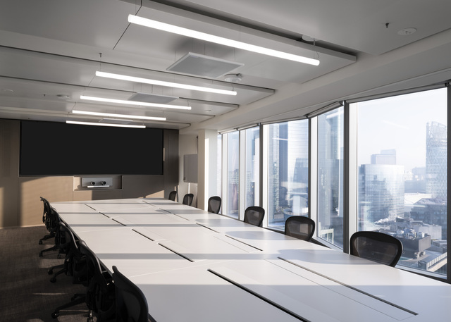 Modern conference room with linear LED ceiling lights illuminating a long white table, black chairs, and floor-to-ceiling city views.