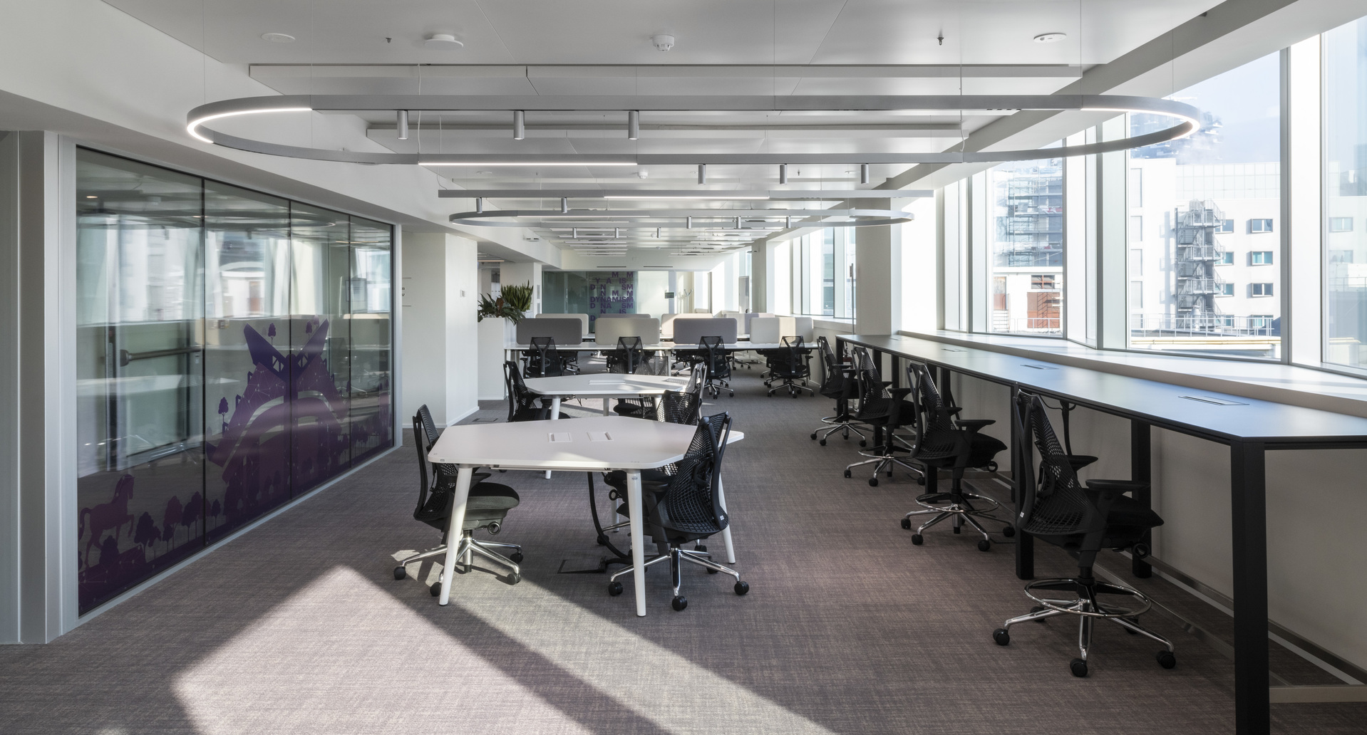 Modern open-plan office with Artemide circular pendant and linear ceiling lights illuminating workstations along floor-to-ceiling windows